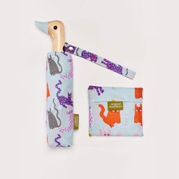 A blue umbrella with a duck head shaped handle next to a folded reusable tote bag on a plain white background